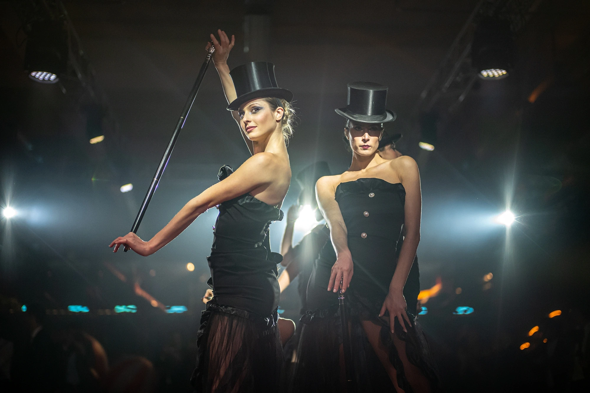 Two performers in top hats and black dresses, canes in hand, under the lights.