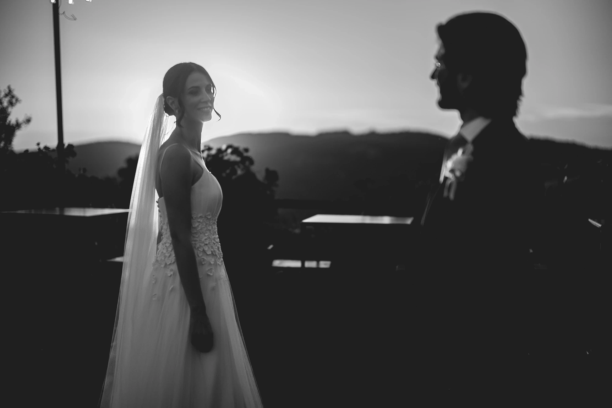 At dusk, the bride in profile and the groom backlit against the hills.
