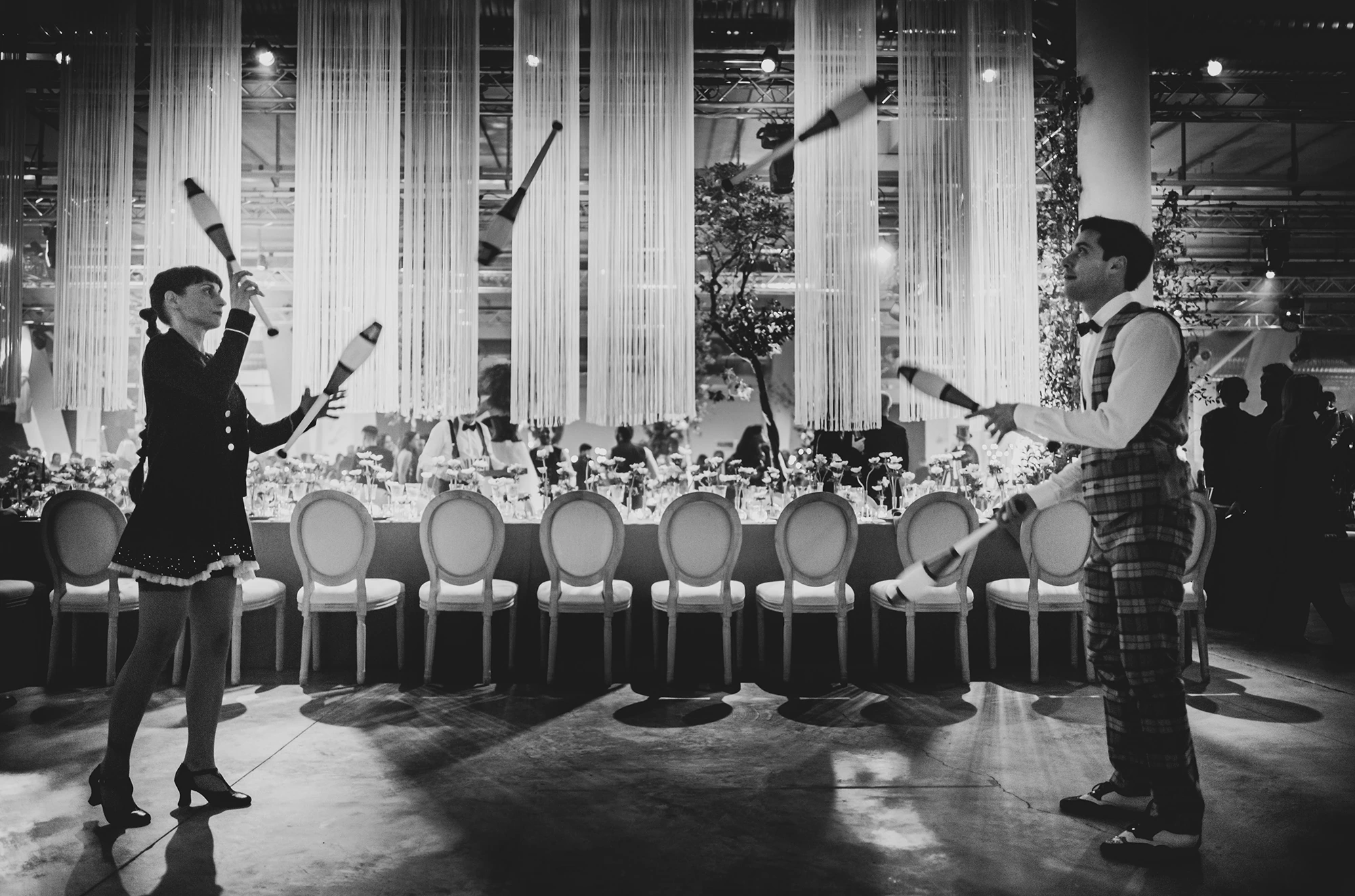 Two jugglers throw clubs along the length of the long table.