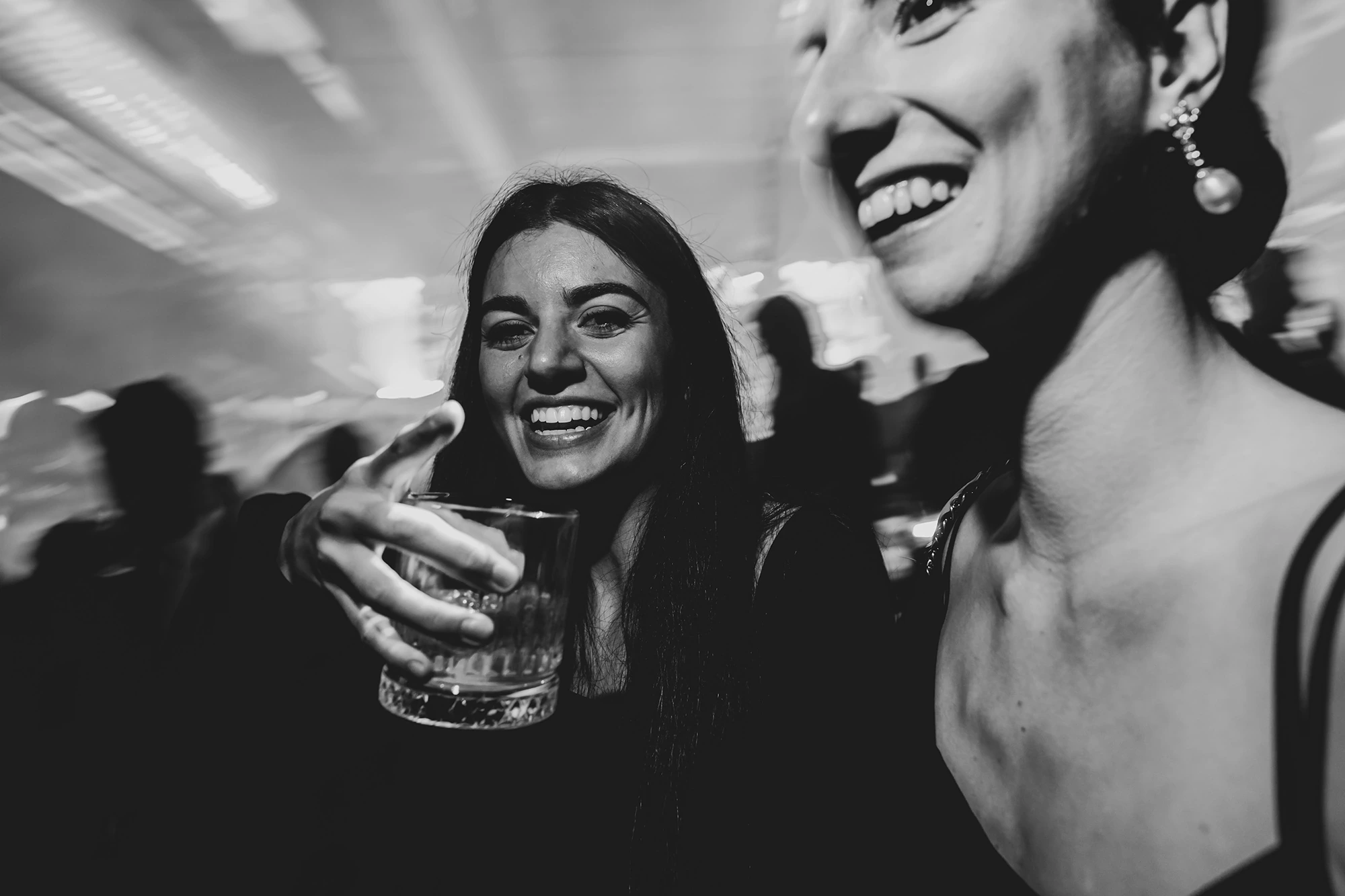 Two women laughing; one raises her glass towards the lens.